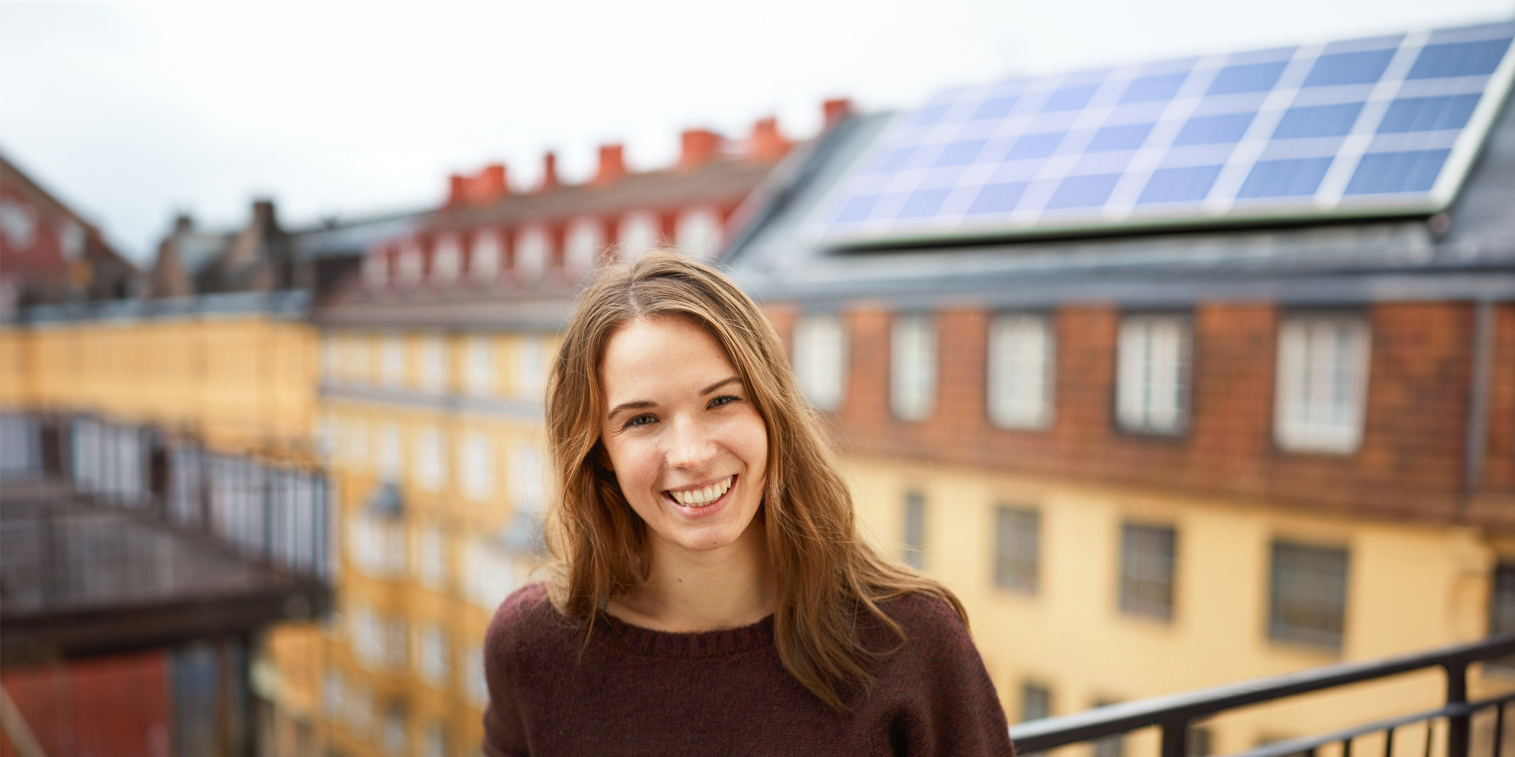 Bild zeigt junge Frau auf einem Balkon, im Hintergrund ein Dach mit Solarpanelen